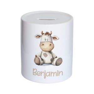 Può includere: Salvadanaio in ceramica bianca con un design di mucca di cartone animato. La mucca è seduta con macchie marroni e ha un naso marrone. Il nome "Benjamin" è scritto in lettere marroni sotto la mucca.