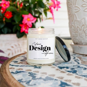 Może przedstawiać: Biała świeca sojowa z czarną i białą etykietą z napisem "Your Design Here" i "Soy Candle Mockup". Świeca stoi na stole z kwiatowym wzorem i zapaloną knot.