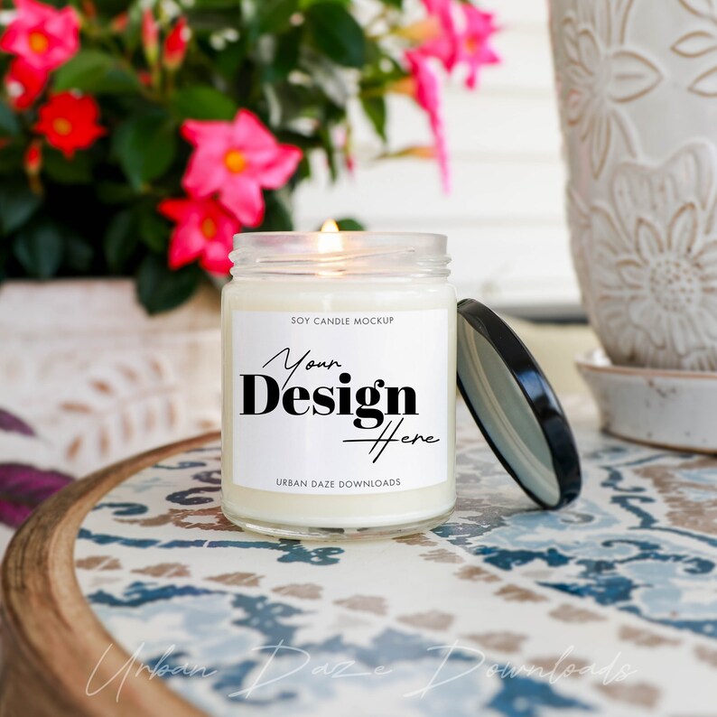 Może przedstawiać: Biała świeca sojowa z czarną i białą etykietą z napisem "Your Design Here" i "Soy Candle Mockup". Świeca stoi na stole z kwiatowym wzorem i zapaloną knot.
