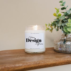 Może przedstawiać: Biała świeca sojowa w szklanym słoiku z białą etykietą, na kt&oacute;rej widnieje napis "Soy Candle Mockup" i "Your Design Here", a na dole tekst "Urban Daze Downloads". Świeca jest zapalona, a obok niej stoi mała zielona roślina w przezroczystym szklanym wazonie.