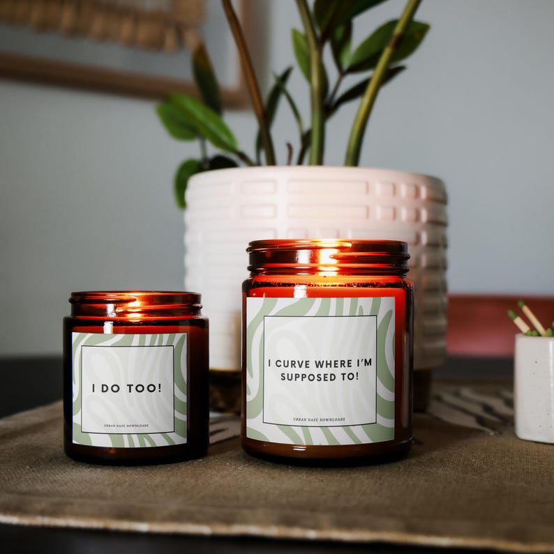 K&ouml;nnte beinhalten: Zwei Kerzenbeh&auml;lter aus bernsteinfarbenem Glas mit wei&szlig;en Etiketten und gr&uuml;nen abstrakten Designs. Der gr&ouml;&szlig;ere Beh&auml;lter tr&auml;gt den Text "I curve where I'm supposed to!" und der kleinere Beh&auml;lter tr&auml;gt den Text "I do too!"