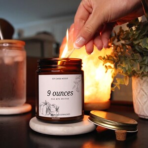 May include: A brown glass candle jar with a white label that says "Soy Candle Mockup" and "9 ounces PSD MP4 Mockup". The candle jar is sitting on a white coaster and has a cork lid.