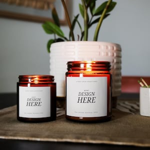 K&ouml;nnte beinhalten: Zwei Kerzenbeh&auml;lter aus bernsteinfarbenem Glas mit wei&szlig;en Etiketten. Die Etiketten tragen den Text "your design here" und "hot candle mockup | PSD".