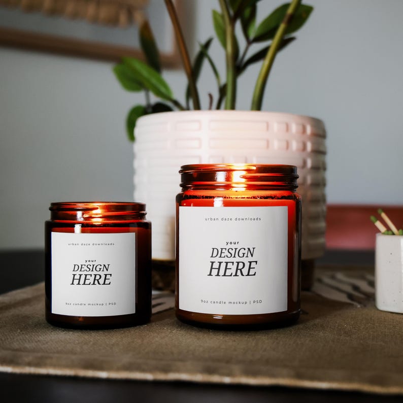 K&ouml;nnte beinhalten: Zwei Kerzenbeh&auml;lter aus bernsteinfarbenem Glas mit wei&szlig;en Etiketten. Die Etiketten tragen den Text "your design here" und "hot candle mockup | PSD".