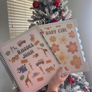 May include: A spiral-bound scrapbook with the name "Rhodes John" and zoo-themed illustrations, including animals and a jeep. The right page says "Baby Girl" with flower designs. The background includes a Christmas tree.