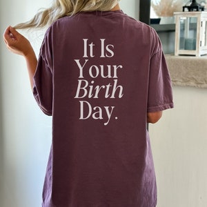 Puede incluir: Una camiseta morada con el texto "It Is Your Birth Day." impreso en blanco en la parte posterior.