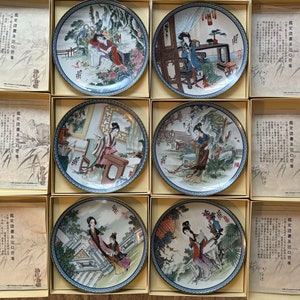Puede incluir: Seis platos decorativos de porcelana con diseños intrincados que representan mujeres con ropa tradicional china. Cada plato está enmarcado en una caja de color crema con una descripción impresa en caracteres chinos.