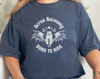 May include: A navy blue t-shirt with a white graphic of a motorcycle with wings, the text "Acrux Raceway" above, and "Born to Ride" below. The shirt is worn by a person wearing blue jeans.