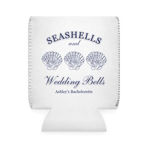 Może przedstawiać: Biały izolator do napoj&oacute;w z napisem "SEASHELLS and Wedding Bells" i trzema ilustracjami muszli w kolorze granatowym. Zawiera r&oacute;wnież tekst "Ashley's Bachelorette".