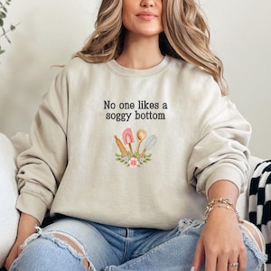 May include: A beige sweatshirt with the text "No one likes a soggy bottom" and a graphic of kitchen utensils with flowers.
