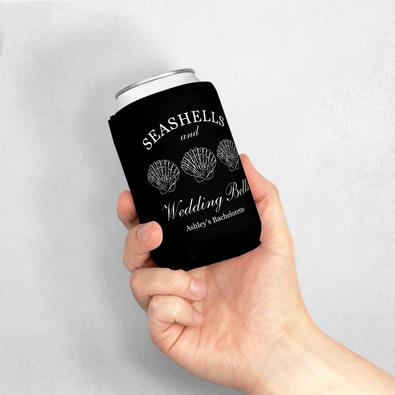 Może przedstawiać: Czarny ochraniacz na puszkę z białym tekstem i grafiką muszli. Tekst brzmi "SEASHELLS and Wedding Bells Ashley's Bachelorette". Ochraniacz ma za zadanie utrzymywać zimne napoje.