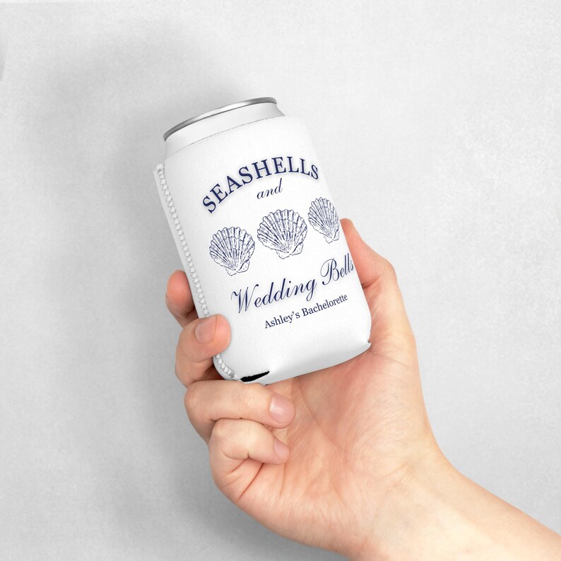 Może przedstawiać: Biała osłona na puszkę z napisem "Seashells and Wedding Bells" i trzema granatowymi ilustracjami muszli. Zawiera r&oacute;wnież tekst "Ashley's Bachelorette". Osłona ma za zadanie utrzymywać zimne napoje.