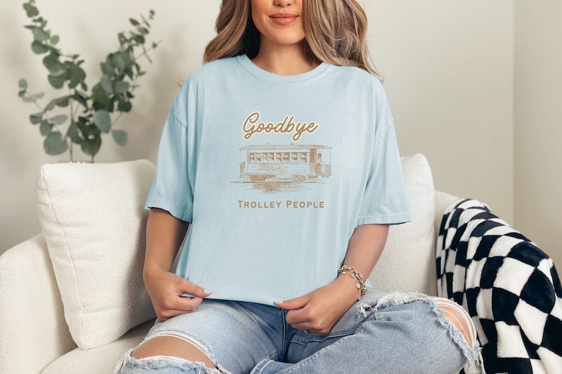 May include: A light blue t-shirt featuring a vintage-style illustration of a trolley car and the text "Goodbye Trolley People".