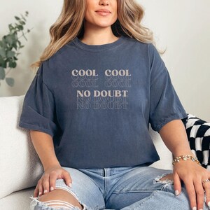 May include: A woman wearing a dark blue oversized t-shirt with the text "COOL COOL COOL COOL NO DOUBT NO DOUBT NO DOUBT" printed on the front. She is sitting on a white couch with a black and white checkered throw blanket.