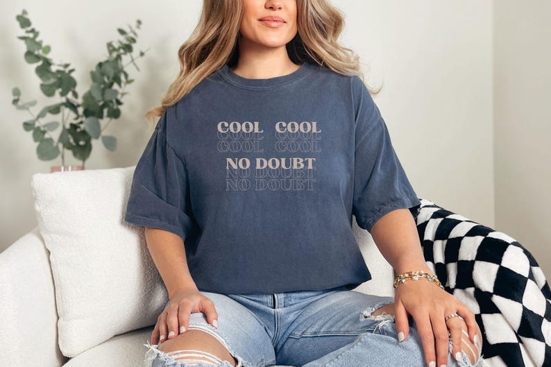 May include: A woman wearing a dark blue oversized t-shirt with the text "COOL COOL COOL COOL NO DOUBT NO DOUBT NO DOUBT" printed on the front. She is sitting on a white couch with a black and white checkered throw blanket.