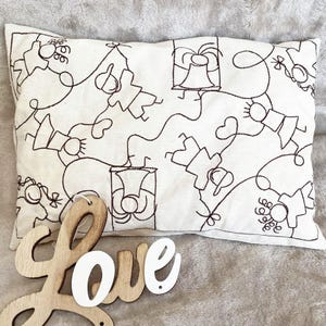 May include: A white pillow with a brown embroidered design of stick figures holding bells. The pillow is sitting on a beige surface with a wooden sign that says "Love" in white letters.