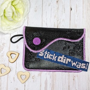 Könnte beinhalten: Eine schwarze Ledertasche mit einem lila Knopf und einem blauen gestickten Aufnäher mit der Aufschrift "stick dir was!" Die Tasche hat einen rosa gestickten Rand und einen schwarzen Kordelzug.