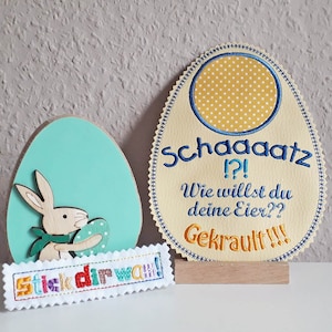 May include: A wooden Easter egg decoration with a bunny holding a smaller egg. The egg has a light blue background and the bunny is brown with a blue bow. The text "Stick dir wan!" is embroidered on a white banner below the egg. A second wooden egg decoration is also shown. This egg is light yellow with a yellow and white polka dot circle in the center. The text "Schagaatz!?! Wie willst du deine Eier?? Gekrault!!!" is embroidered on the egg in blue and yellow.
