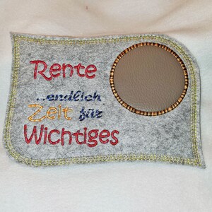 Puede incluir: Un letrero de fieltro gris con un borde cosido amarillo. El letrero tiene un c&iacute;rculo marr&oacute;n en el centro con un fondo gris. El texto en el letrero dice "Rente...endlich Zeit f&uuml;r Wichtiges" en bordado rojo, amarillo y azul.