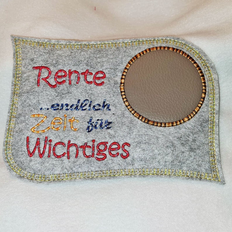 Puede incluir: Un letrero de fieltro gris con un borde cosido amarillo. El letrero tiene un c&iacute;rculo marr&oacute;n en el centro con un fondo gris. El texto en el letrero dice "Rente...endlich Zeit f&uuml;r Wichtiges" en bordado rojo, amarillo y azul.
