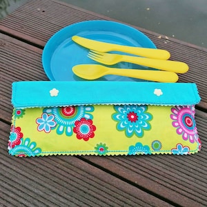 May include: A turquoise fabric cutlery holder with a floral pattern and white buttons. The holder is folded and sitting on a wooden surface. Three yellow plastic utensils are sitting on a blue plate behind the holder.