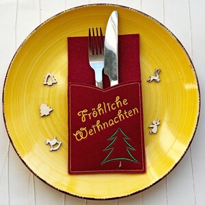 May include: A yellow ceramic plate with a red felt cutlery holder featuring the text "Fröhliche Weihnachten" and a green Christmas tree design. A fork and knife are in the holder. Small wooden Christmas ornaments are scattered around.