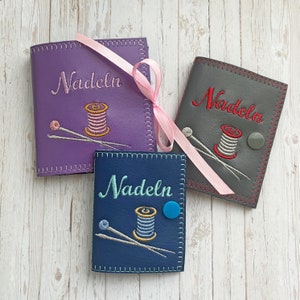 May include: Three purple, gray, and blue needle cases with the word "Nadeln" embroidered on each. Each case features a spool of thread and two needles.