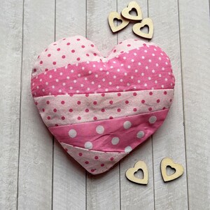Könnte beinhalten: Ein rosa herzförmiges Kissen mit weißen Tupfen und Streifen. Kleine Holzherz-Dekorationen sind darum herum verstreut. Das Kissen besteht aus Patchwork-Stoff mit verschiedenen Rosa- und Weißtönen.