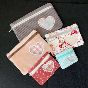 Puede incluir: Una colección de neceseres con cremallera en varios tamaños y colores. Los neceseres presentan recortes en forma de corazón con diferentes telas estampadas visibles a través de las aberturas. Los colores incluyen gris, rosa, rojo, floral y verde menta. Cada neceser tiene una cremallera en contraste.