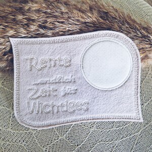 Puede incluir: Un parche de fieltro blanco con el texto "Rente endlich Zeit fur Wichtiges" bordado en &eacute;l. El parche tiene un c&iacute;rculo en el centro.