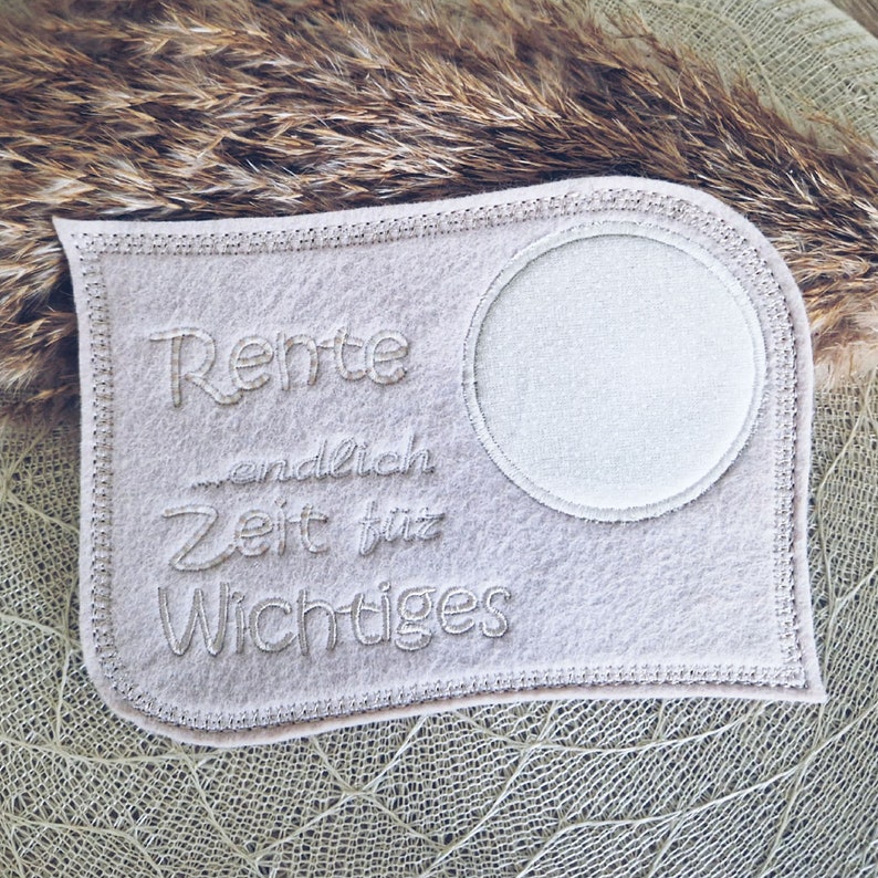 Puede incluir: Un parche de fieltro blanco con el texto "Rente endlich Zeit fur Wichtiges" bordado en &eacute;l. El parche tiene un c&iacute;rculo en el centro.