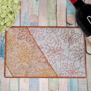 May include: A rectangular beige coaster with an embroidered design of brown and red leaves. The coaster has a brown border.