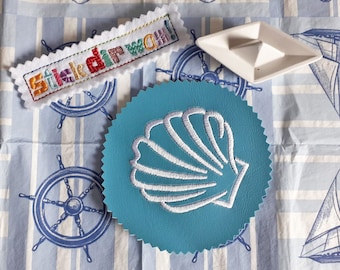 May include: A blue and white embroidered seashell patch with the text "Stickd'rwach!" on a white and blue patterned fabric background.
