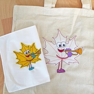 May include: Two embroidered tote bags with a cartoon maple leaf design. One leaf is yellow and the other is white. Both leaves are playing a trumpet.