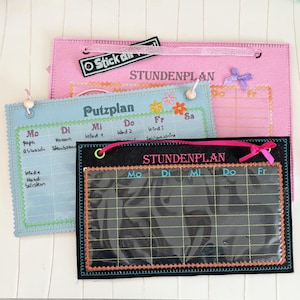 May include: Three felt boards with different designs. The top board is pink with the text "STUNDENPLAN" and a small pink bow. The middle board is blue with the text "Putzplan" and a floral design. The bottom board is black with the text "STUNDENPLAN" and a pink bow. All three boards have a clear plastic window for writing.