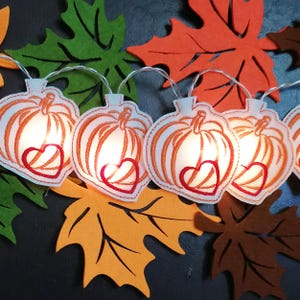 Könnte beinhalten: Eine Lichterkette mit Kürbis-förmigen Ornamenten, orangefarbenen Konturen und einem roten Herzdesign. Filzblätter in Orange, Grün und Braun ergänzen das herbstliche Thema und die Dekoration.