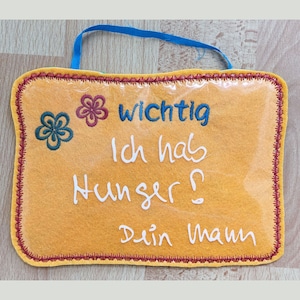 May include: A rectangular, yellow felt sign with a blue ribbon for hanging. The sign has embroidered text in German that reads "wichtig Ich hab Hunger? Dein Mann" and is decorated with embroidered flowers. The edges are trimmed with red stitching.