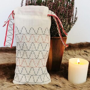May include: A white fabric drawstring bag with a red and black trim. The bag is decorated with a repeating pattern of embroidered pine trees. The bag is sitting on a brown furry surface next to a lit white candle.