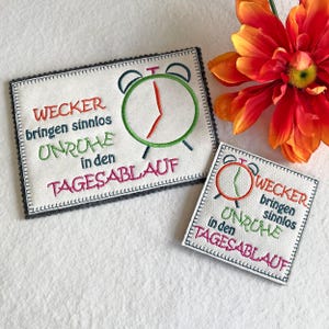 May include: Two embroidered fabric squares with a white background and gray border. Each square features a red and green alarm clock with the text "Wecker bringen sinnlos Unruhe in den Tagesablauf" in colorful embroidery.