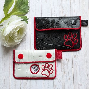 Pode incluir: Duas bolsas de couro preto com zíper e costuras vermelhas. A bolsa maior tem um design floral e uma pegada de pata. A bolsa menor tem uma pegada de pata e um círculo com um design dentro. Ambas as bolsas têm botões vermelhos.