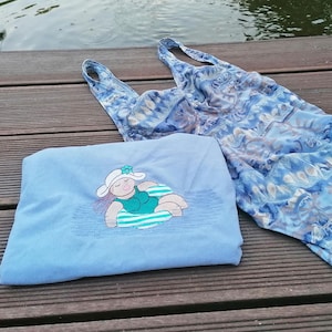 May include: Two swimsuits and a light blue towel on a wooden dock. One-piece swimsuit with a blue and white pattern. The other swimsuit has straps and a similar pattern. The towel has an embroidered design of a person in a swimsuit floating on a tube.