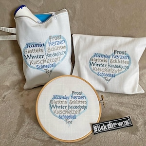 May include: A collection of white fabric items with blue embroidered text. The items include a zippered pouch, a pillow, and an embroidery hoop. The text is arranged in a heart shape and includes German words like "Frost" and "Winter". A small black patch with white text is also present.