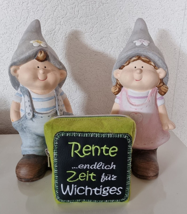 Puede incluir: Una bolsa de fieltro verde con cierre de cremallera y un parche de fieltro negro que dice "Rente ...endlich Zeit f&uuml;r Wichtiges". Dos peque&ntilde;os gnomos grises con caras pintadas est&aacute;n detr&aacute;s de la bolsa.