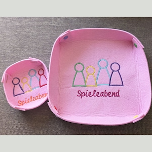 Könnte beinhalten: Zwei rosa Filzschalen mit gestickten Spielfiguren und dem Text "Spielabend". Die kleinere Schale hat vier Spielfiguren und die größere Schale hat fünf Spielfiguren.