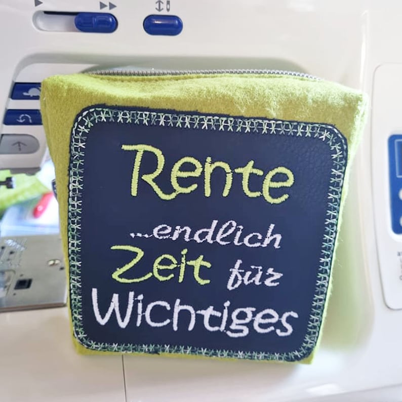 Puede incluir: Una bolsa verde con cremallera con un parche bordado en blanco y negro que dice "Rente...endlich Zeit f&uuml;r Wichtiges".