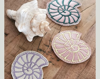 May include: Three embroidered fabric nautilus shell shapes. One is purple, one is pink, and one is green. The shells are on a wooden surface.