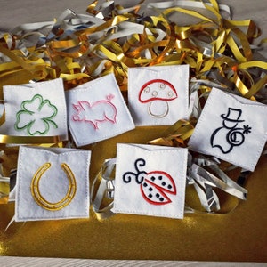 May include: Six small white felt pouches with different embroidered designs. The designs include a four-leaf clover, a pig, a mushroom, a ladybug, a horseshoe, and a snowman.