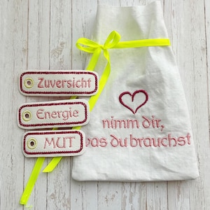 Könnte beinhalten: Eine weiße Stofftasche mit einem gelben Band, das um sie herum gebunden ist. Die Tasche hat eine rosa Stickerei mit der Aufschrift "nimm dir, was du brauchst" und ein rosa Herz. Neben der Tasche liegen drei weiße Stoffanhänger mit roten Rändern und rosa Stickerei. Die Anhänger sagen "Zuversicht", "Energie" und "Mut".