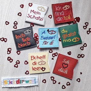 May include: Seven colorful felt squares with different love messages in German. The squares are decorated with red hearts and are scattered on a wooden surface with red heart confetti.