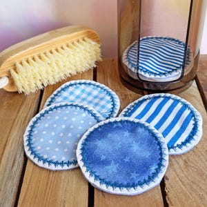 Op de afbeelding: Herbruikbare katoenen gezichtsreinigingspads in verschillende blauwe patronen, waaronder stippen, strepen en sterren, met decoratieve stiksels. Een bamboe borstel en een doorzichtige container zijn ook zichtbaar.